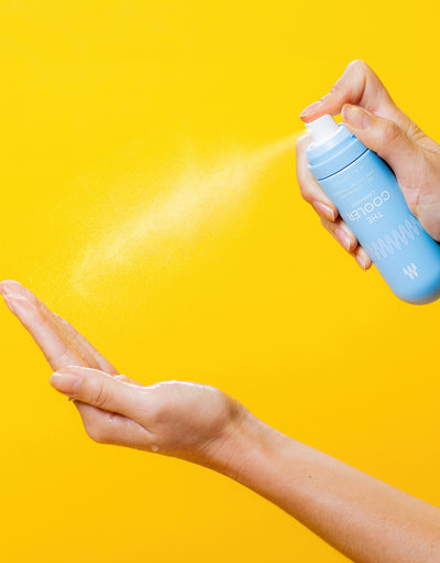Close-up of hands spraying a blue grooming product bottle labeled "THE COOLER" against a bright yellow background.