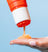 Close-up of orange exfoliating scrub being dispensed onto a hand with a French manicure against a light blue background.