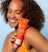 Smiling woman with curly hair holding an orange tube of Meridian's "The Scrub" grooming exfoliator against a light blue background.