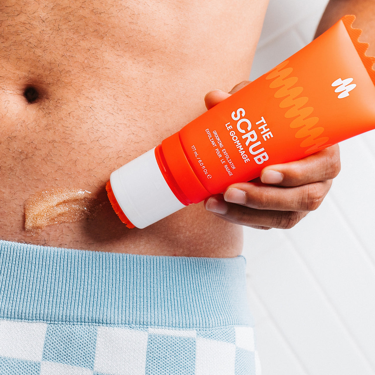 Close-up of exfoliating scrub cream being applied to lower abdomen with orange tube labeled "The Scrub Le Gommage" for skincare.