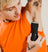 Person wearing orange sleeveless shirt using black electric trimmer to shave underarm hair with visible tattoos on arm and hand