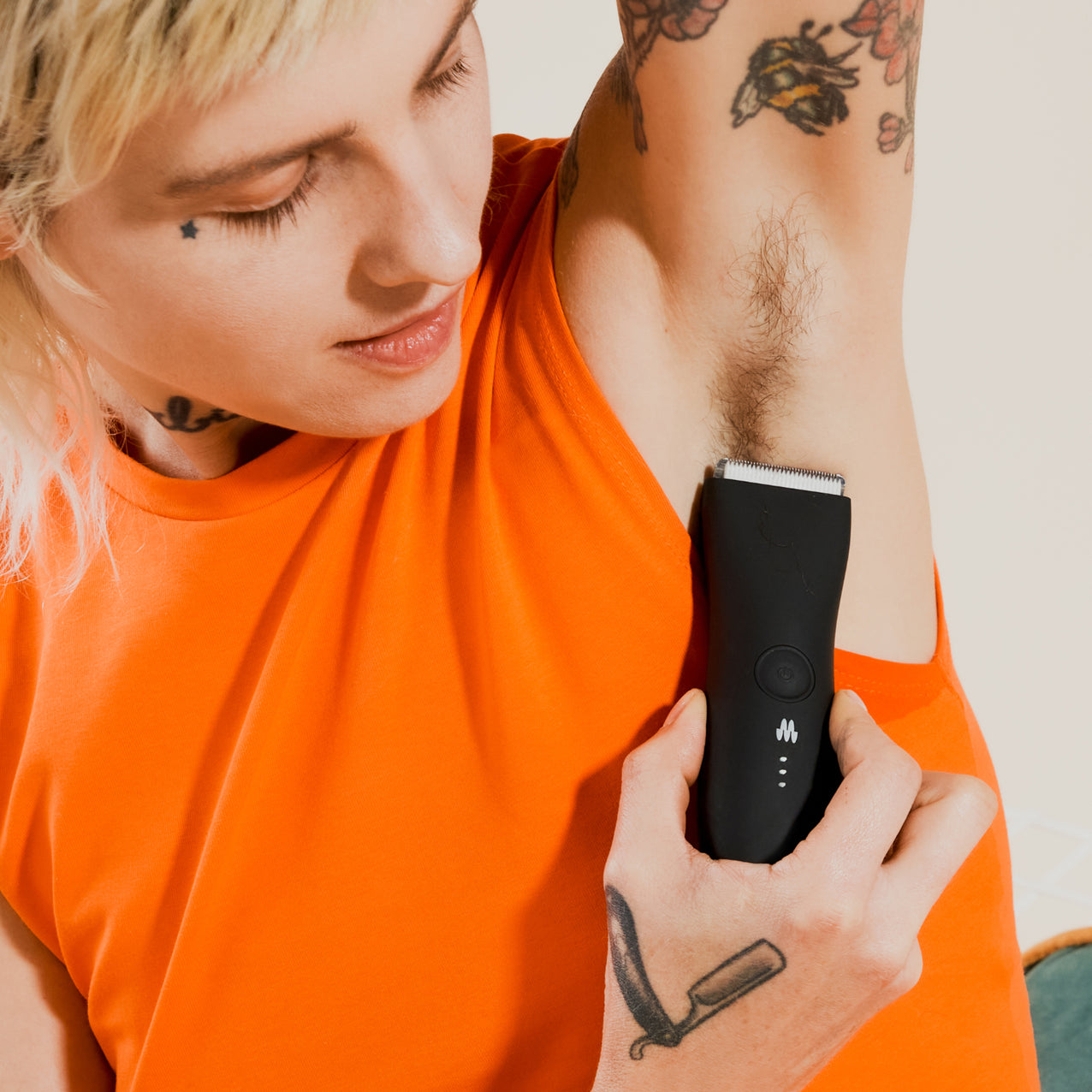 Person wearing orange sleeveless shirt using black electric trimmer to shave underarm hair with visible tattoos on arm and hand
