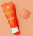 Orange tube of "The Scrub" exfoliating face wash with a translucent orange cap on a peach background, skincare product.