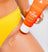Exfoliating body scrub being applied on thigh, bright orange tube labeled "The Scrub," woman in yellow bikini bottom