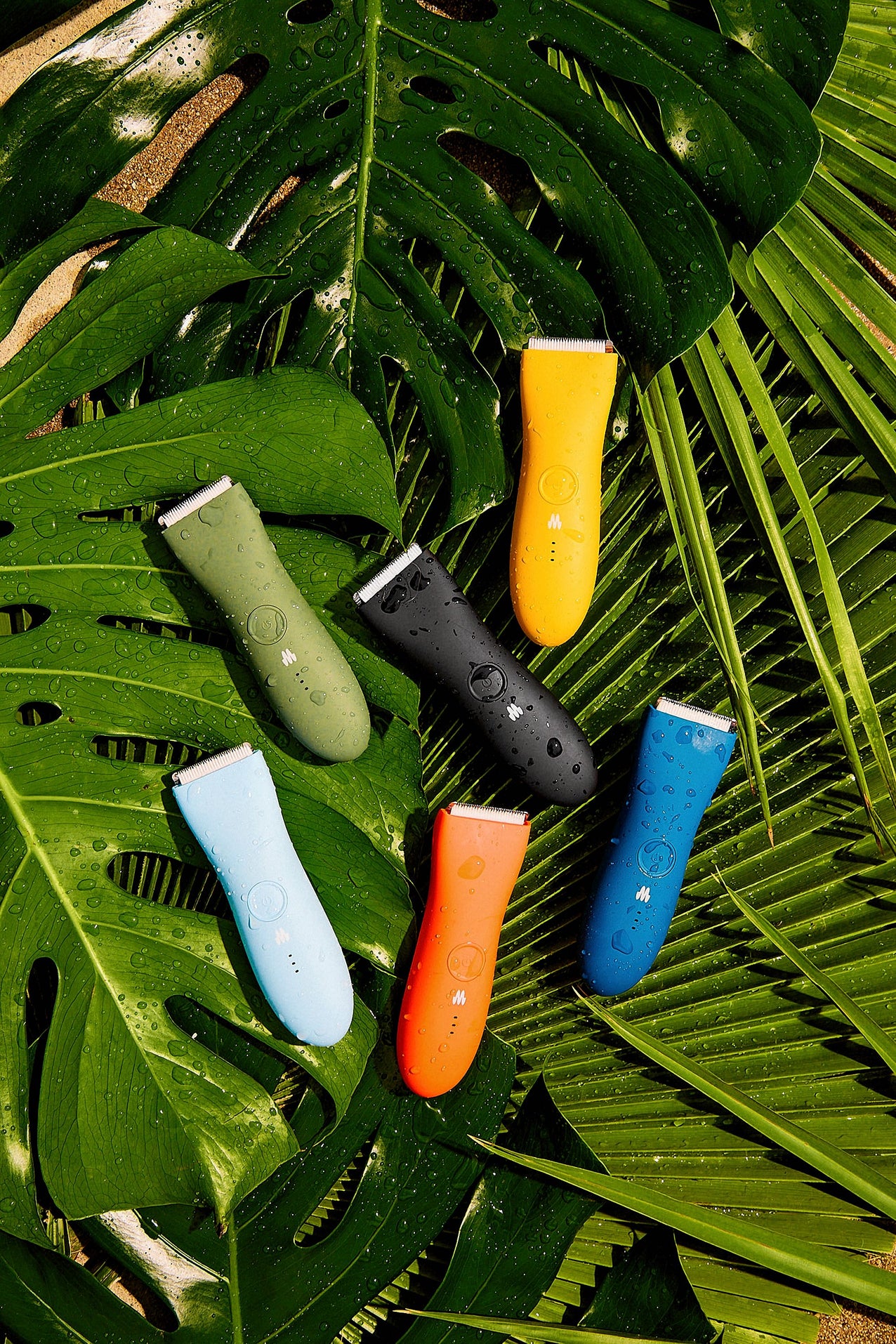 Six colorful water-resistant electric grooming trimmers arranged on large green tropical leaves with water droplets.