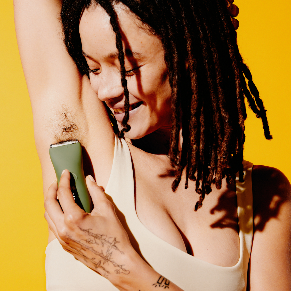 Woman with dreadlocks shaving armpit hair with green electric trimmer against vibrant yellow background, tattooed hand visible.