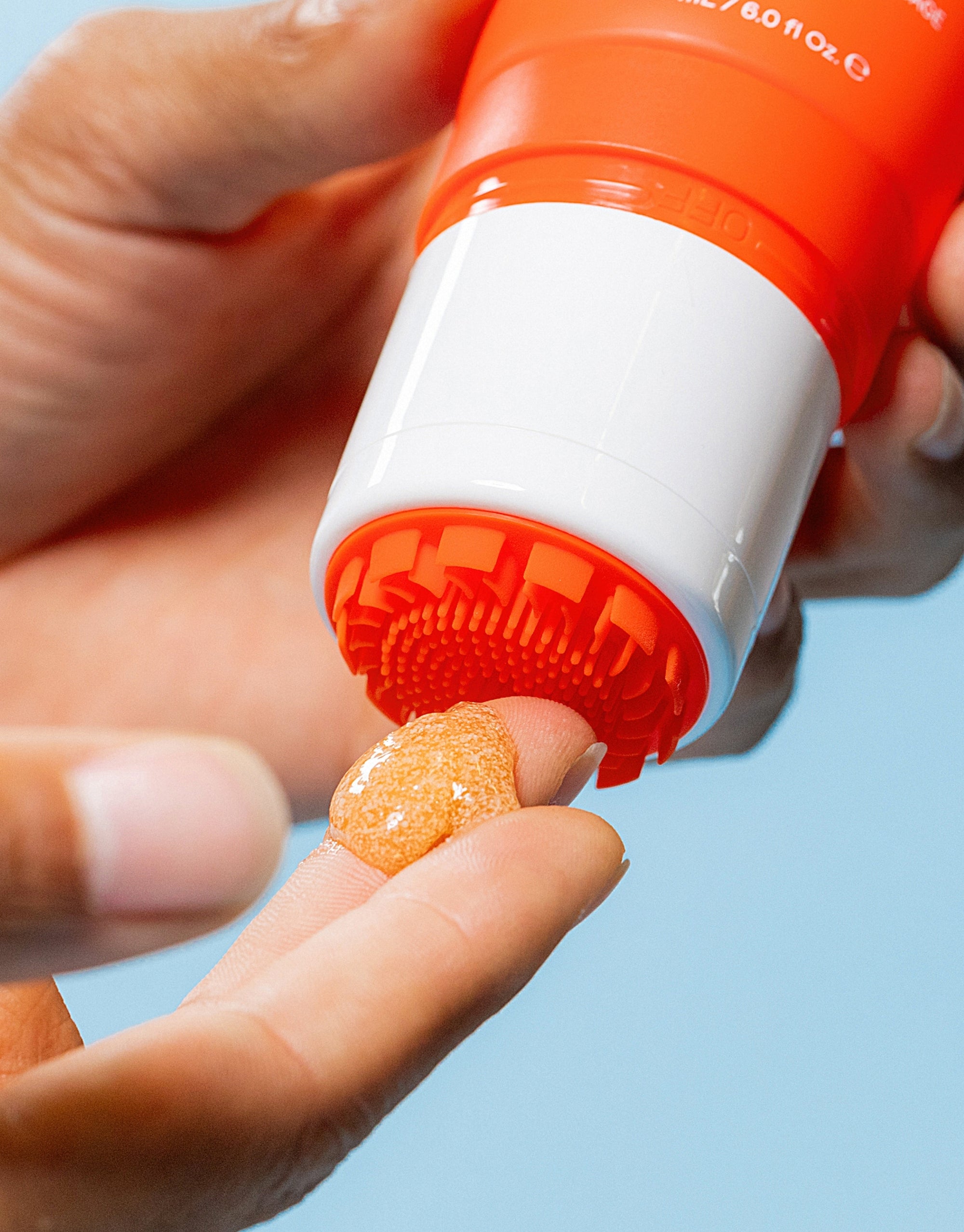 Close-up of a hand dispensing orange exfoliating scrub gel from a red and white tube onto a fingertip.