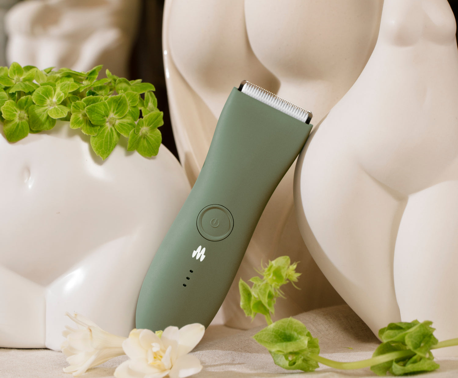 Green rechargeable electric hair clipper with blade guard, placed near white ceramic vases and green leaves on beige surface.