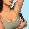 Woman using black electric trimmer to shave armpit hair, wearing green tank top and gold necklaces on light blue background.