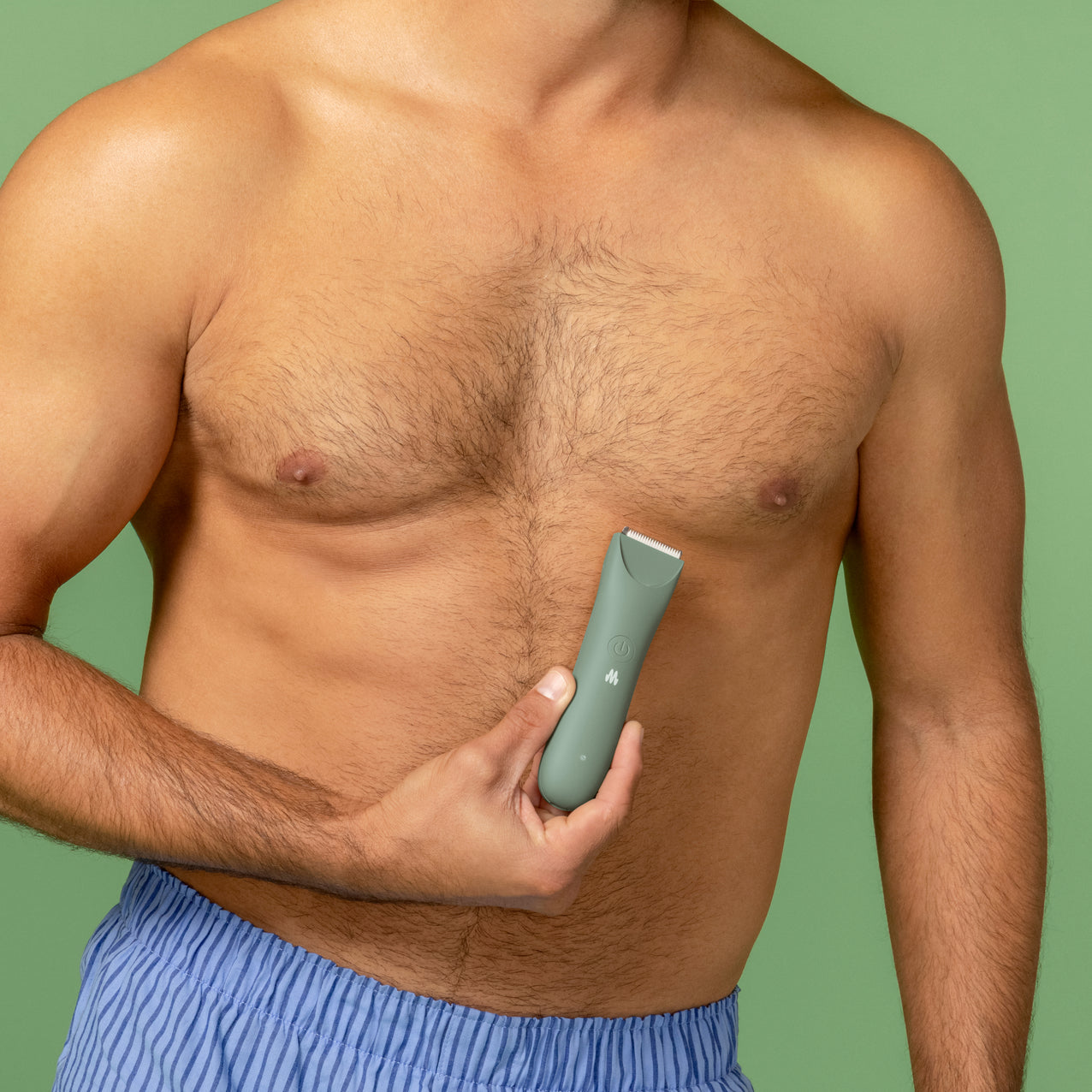 Man with chest hair holding green electric body hair trimmer against bare chest, grooming and personal care tool.
