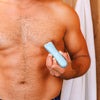Man grooming chest hair with blue electric body hair trimmer in wooden bathroom with white towel hanging.