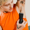Person wearing orange sleeveless shirt using black electric trimmer to shave underarm hair with visible tattoos on arm and hand