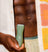 Handheld green facial cleansing brush on dark skin with open beige shirt and patterned orange towel, skincare routine tool.