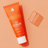 Orange tube of "The Scrub" exfoliating face wash with a translucent orange cap on a peach background, skincare product.