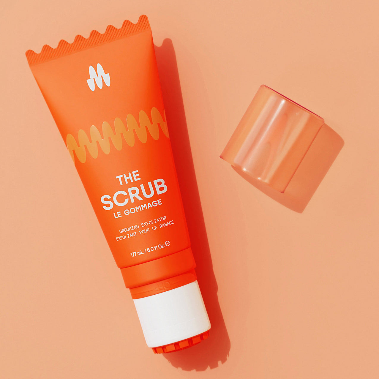 Orange tube of "The Scrub" exfoliating face wash with a translucent orange cap on a peach background, skincare product.