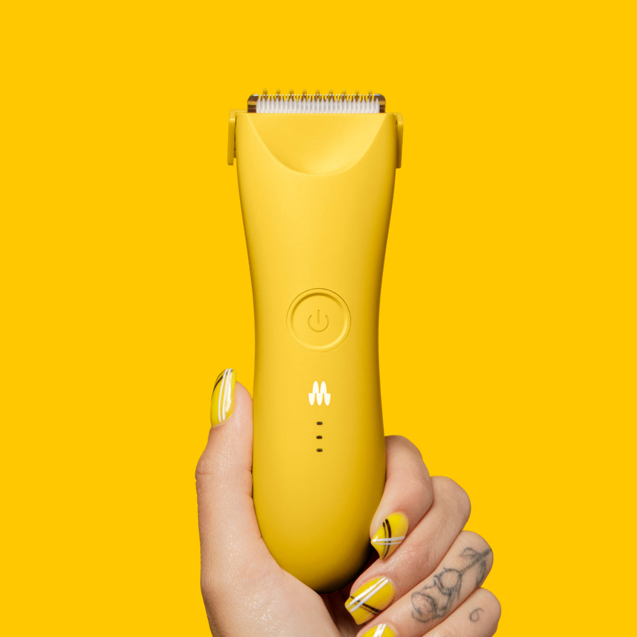 Yellow cordless electric hair clipper with power button held by hand with yellow and black nail art and floral tattoo on fingers.