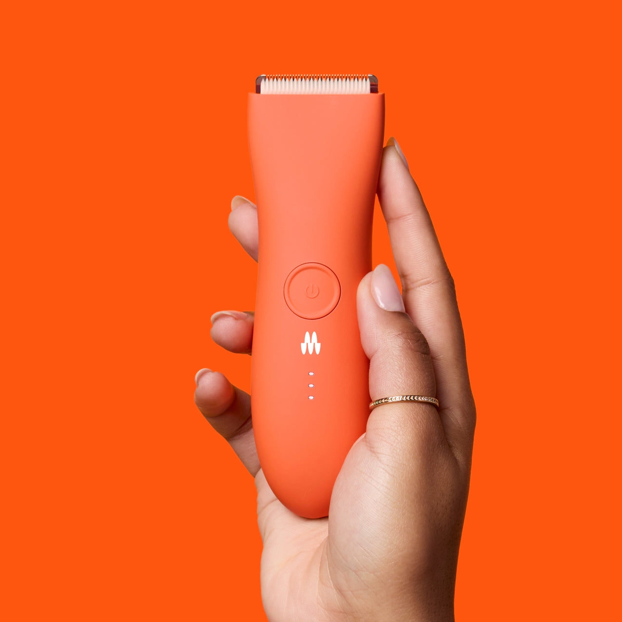 Hand holding a sleek orange cordless hair clipper with power button and LED battery indicators on a matching orange background.