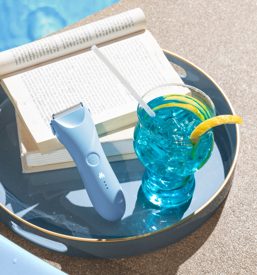 Blue electric shaver, open book, and blue cocktail with lemon on round glass table in bright sunlight